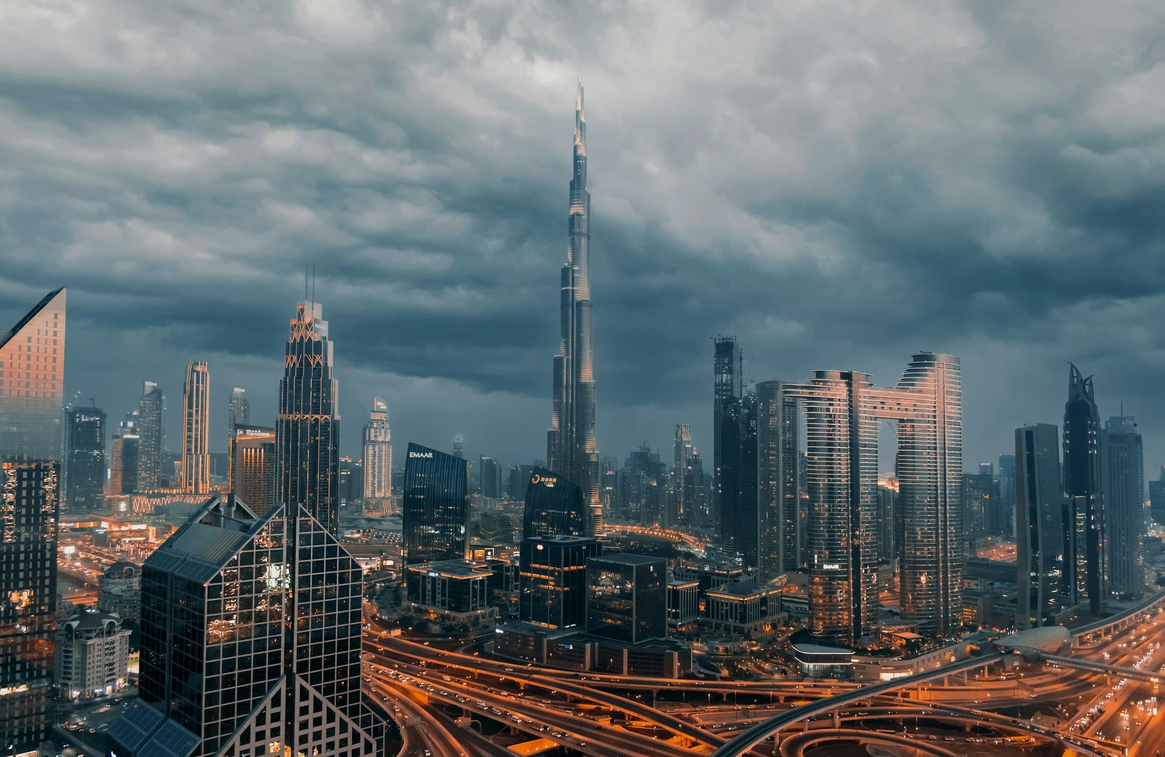 Downtown Dubai at dusk under a dark sky, the Burj Khalifa rising above the surrounding towers and the Sheikh Zayed Road interchange